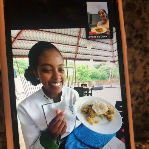 Pantalla de un móvil mostrando a la Chef Gaby dando una clase virtual por Zoom, con una alumna sonriendo y mostrando su plato